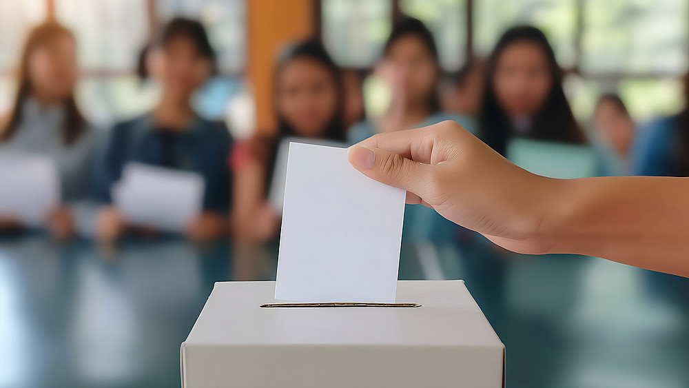 Jugendliche sitzen im Hörsaal im Hintergrund, vor Ihnen sieht man eine Hand, die einen weißen Zettel in eine Wahlurne einwirft. 