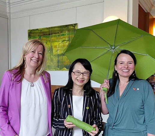 Eine Vertretung aus Taiwan hält einen grünen kompakten Regenschirm in der Hand und steht zwischen der Prorektorin und Frau Fentzel in einem Sitzungsraum. Frau Fentzel hält einen grünen Regenschirm aufgespannt über ihrem Kopf.