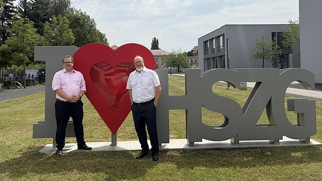 : Prof. Dr.-Ing. Alexander Kratzsch und Prof. Dr.-Ing. Sascha Müller-Feuerstein vor dem I Love HSZG Logo auf dem Campus