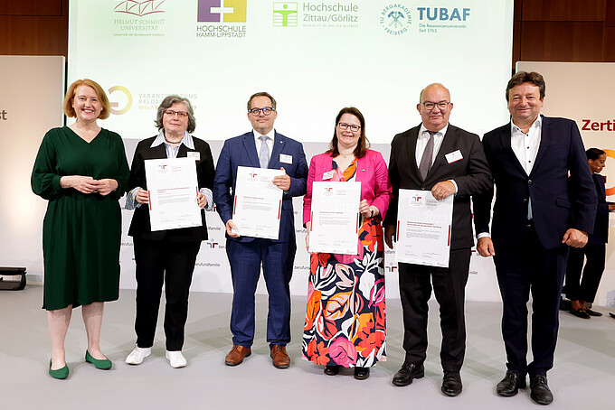 Die Bundesministerin für Familie und Vertretende der zertifizierten Hochschulen stehen auf der Bühne und halten ihre Zertifikate in den Händen.