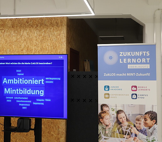 Ein Bildschirm mit einer Präsentation steht vor einer Korkwand, daneben ein ZUKLOS-Rollup.