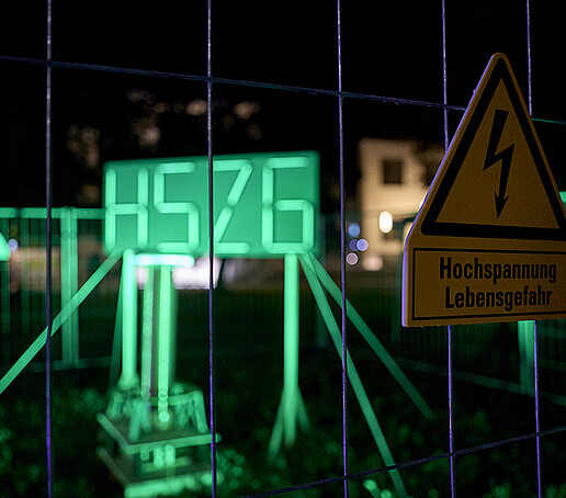 Das HSZG-Logo leuchtet in neongrün durch Leuchtstoffröhren.