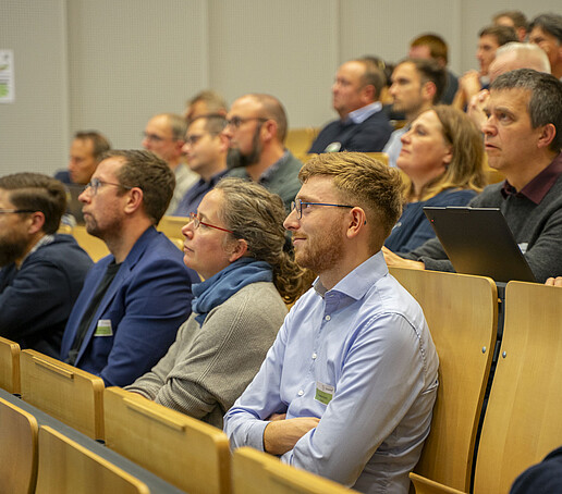 Die Sitzreihen sind mit Publikum besetzt, dass den Blick nach vorn richtet.