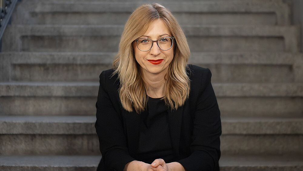 Prof. Dr. rer. pol. Jana Stolz-Römmermann trägt einen schwarzen Blazer und eine Brille, sitzt auf einer Treppenstufe und lächelt in die Kamera.