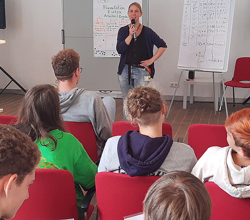Eine Gruppe von Schülern sitzt auf Stühlen vor einer Sprecherin, die das Camp moderiert. 
