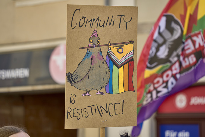 Ein Schild mit der Aufschrift: Community is Resistance wird in einer Straßenmenge hochgehalten, eine Regenbogenflagge weht daneben.