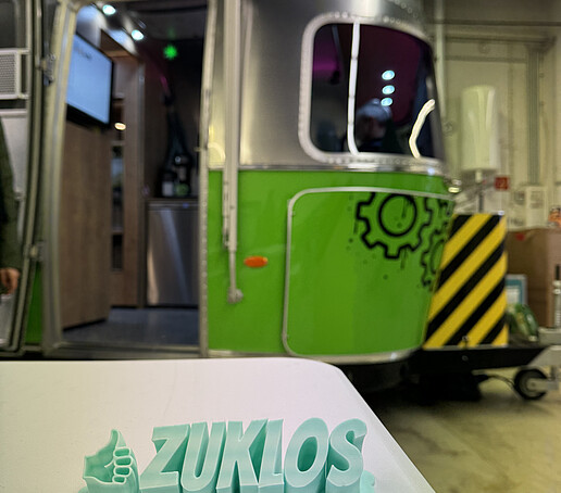 Vor dem AirStream-Anhänger liegt ein 3D-Ausdruck des Schriftzuges ZUKLOS auf einem Tisch.