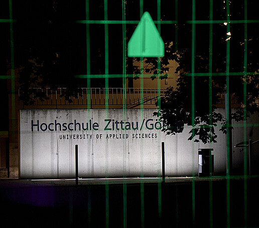 Der Schriftzug Hochschule Zittau/Görlitz leuchtet hell, im Vordergrund ist ein grün erleuchteter Bauzaun.