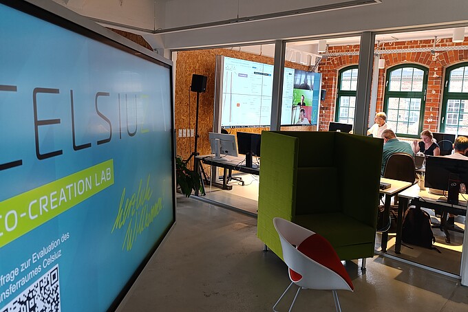 Das Celsiuz-Logo auf einem Bildschirm steht im Vordergrund eines Arbeitsraumes, in dem Menschen an Tischen sitzen und auf einen Großbildschirm schauen.