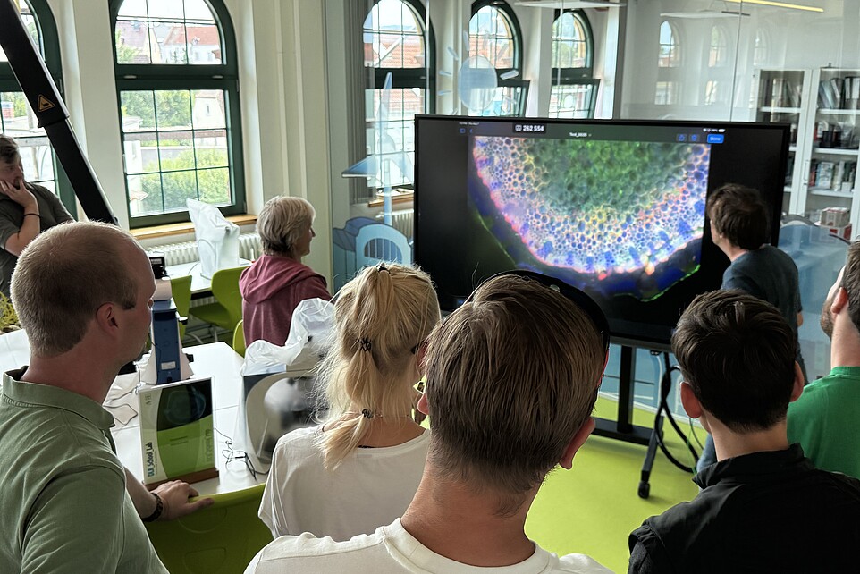 Lehrer und Lehrerinnen stehen im Schülerlabor vor Versuchsständen und blicken auf einen Bildschirm.