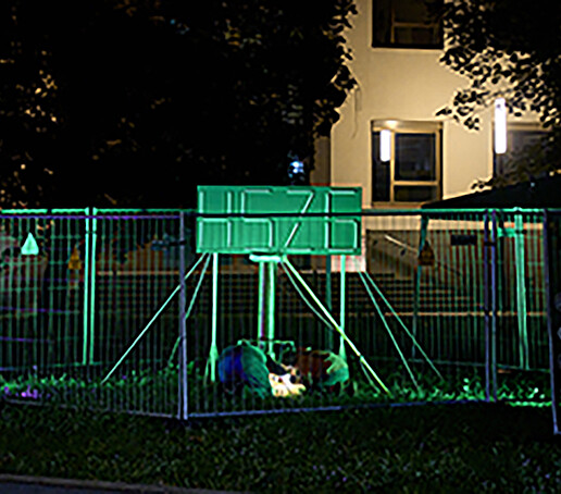 Eine grünleuchtende Lichtinstallation in Form des HSZG-Logos steht auf der Wiese vor dem HSZG-Gebäude umgeben von Bauzäunen.