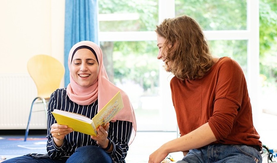 Zwei junge Frauen sitzen in einem Seminarsaal auf dem Boden, die linke Frau hält ein aufgeschlagenes Buch in der Hand. Beide lachen.