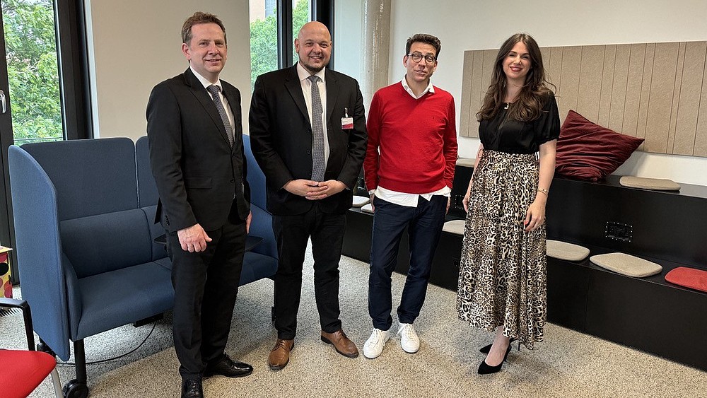 Four people stand in a room in front of chairs and look into the camera.