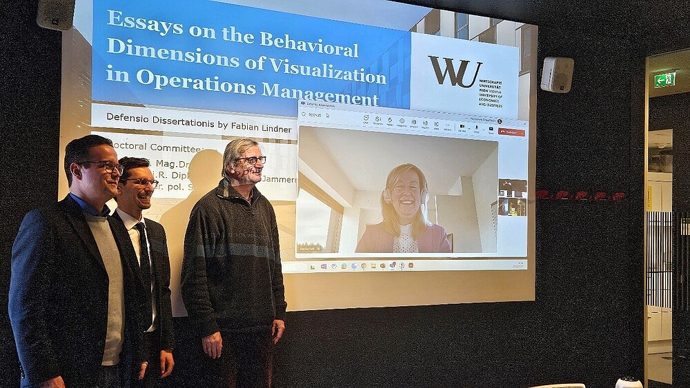 Drei Männer stehen links vor einer erleuchteten Beamerleinwand, auf der Professorin Keil zu sehen ist.