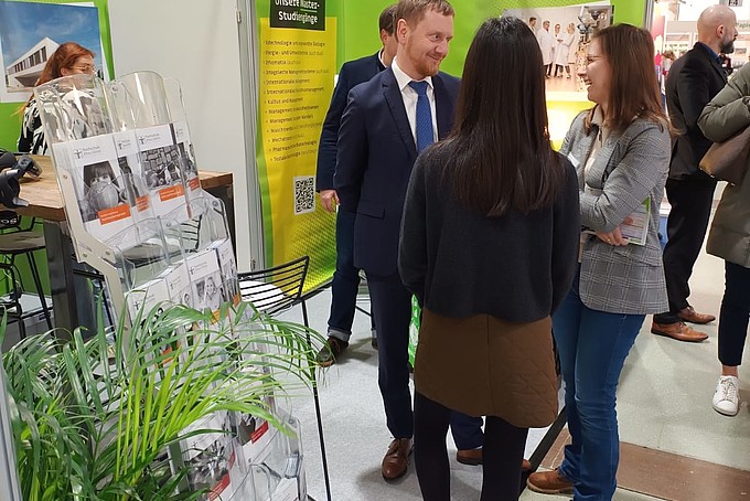 Ministerpräsident Kretschmer, die Studienberaterin der HSZG und Zhaniya unterhalten sich am Messestand.