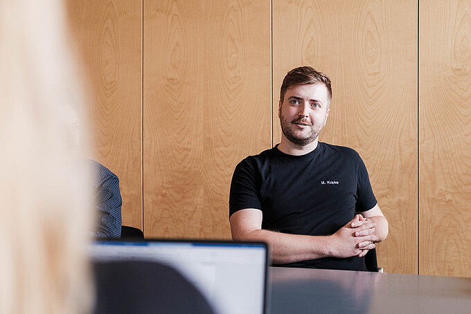 Ein Mann im schwarzen T-Shirt sitzt an einem Tisch und schaut in die Gesprächsrunde.
