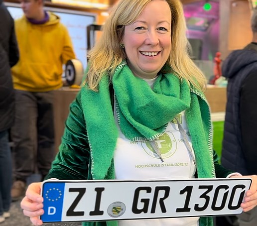 Die Prorektorin hält das Sprinter-KfZ-Kennzeichen in den Händen und steht vor dem geöffneten AirStream-Anhänger.