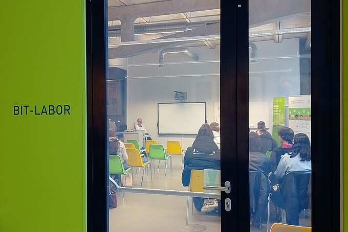Durch eine verschlossene Glastür sieht man Stuhlreihen von hinten, auf denen Schülerinnen und Schüler sitzen.