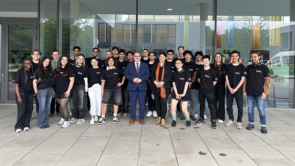 Eine Gruppe ausländischer Studierender und der Rektor stehen vor der Glasfassade von Haus Z vier und lächeln in die Kamera.