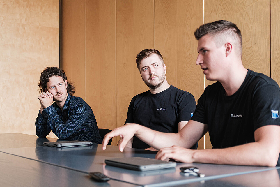 Drei KIA-Studenten sitzen an einem Tisch bei einer Besprechung vor einer holzvertäfelten Wand.