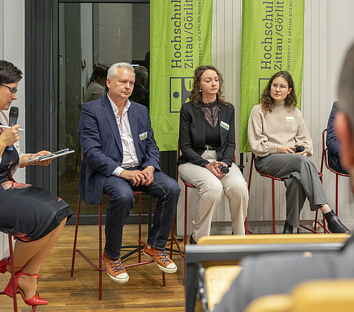 Im Panel sitzen Talk-Gäste auf Hochstühlen nebeneinander, die Moderatorin spricht in ein Mikrofon.