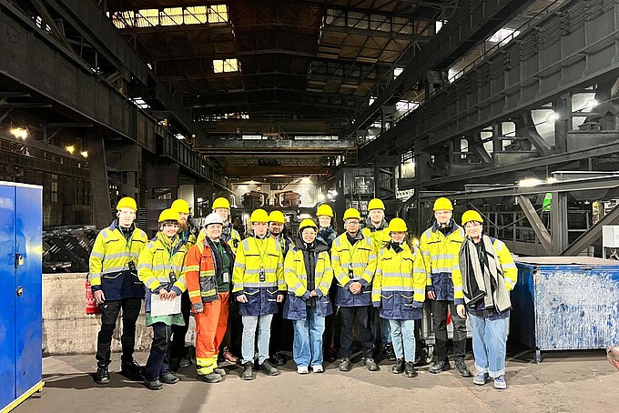 Eine Gruppe Studierender in gelber Schutzkleidung mit gelben Helmen steht in einer Fabrikhalle und lächelt in die Kamera.